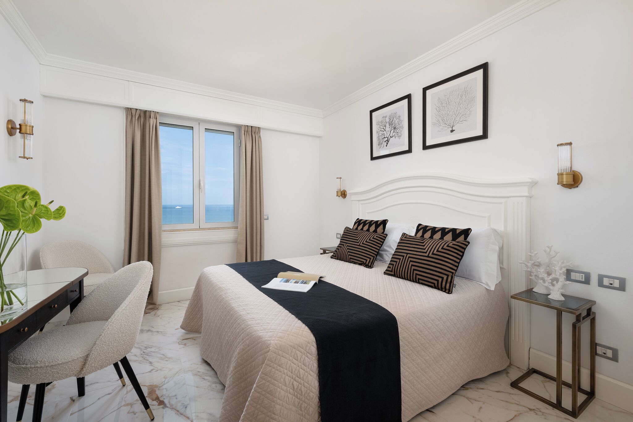 A hotel room with a large bed, a desk with a chair, a small table, and a view of the sea through the window.