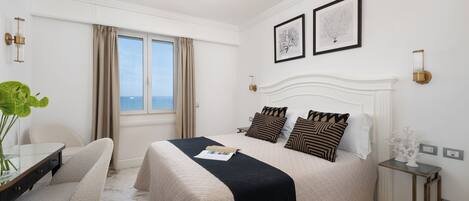 A hotel room with a large bed, a desk with a chair, a small table, and a view of the sea through the window.