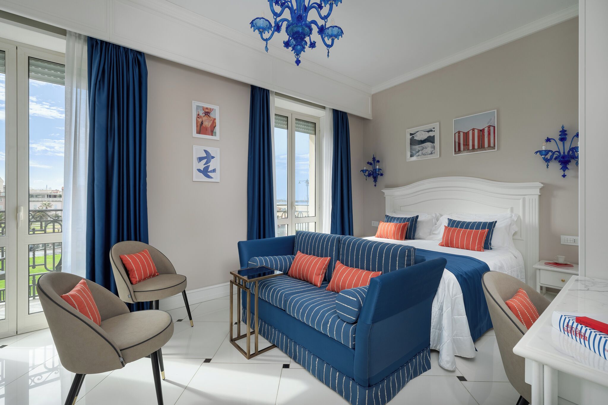 Habitación de hotel con cama, sofá azul a rayas, una silla, una mesita y vistas al exterior.