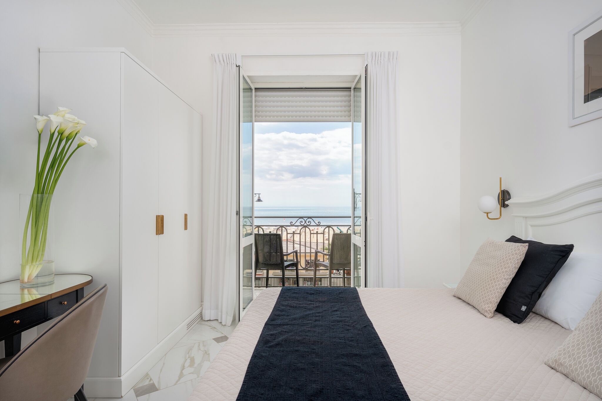 Et hotelværelse med seng, skrivebord, blomstervase, balkon med havudsigt og et vindue med gardiner.