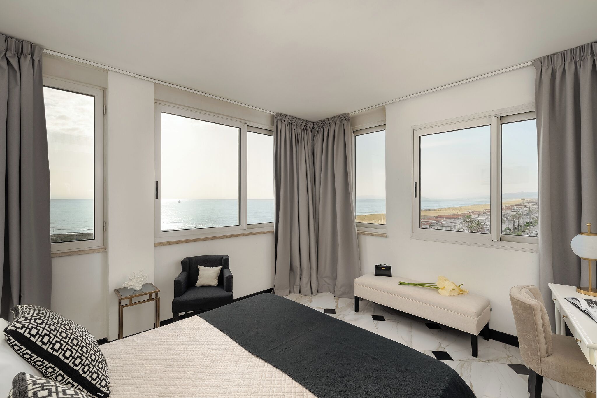 A hotel room with a bed, a chair, a bench, a desk, and large windows with curtains.