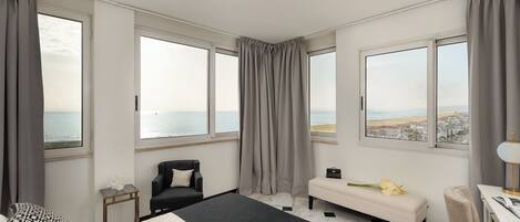 A hotel room with a bed, a chair, a bench, a desk, and large windows with curtains.