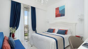 Habitación de hotel con cama, escritorio, silla, ventana con cortinas, candelabro y un cuadro en la pared.