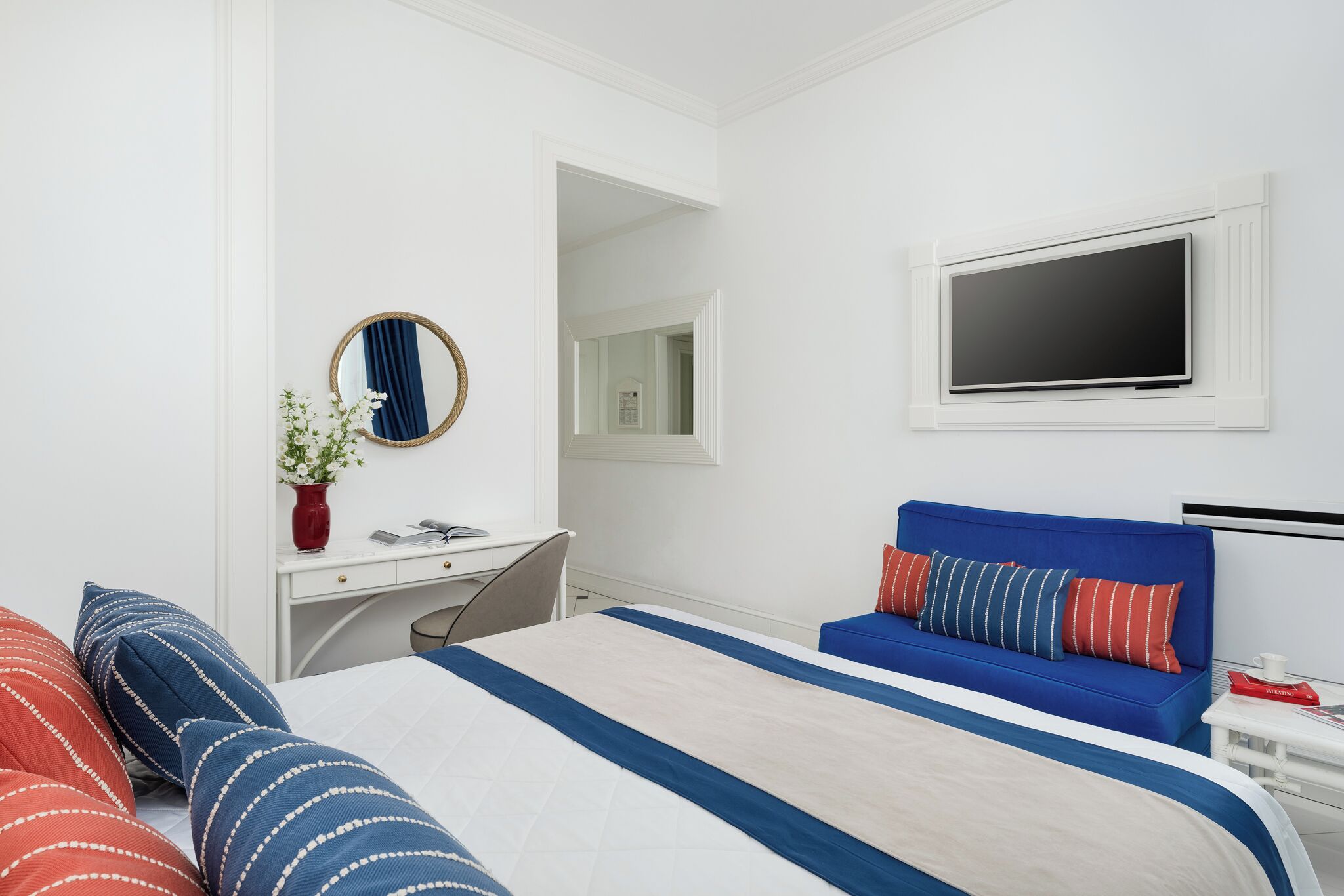 A modern hotel room with a bed, a desk, a blue sofa, a TV, and a mirror.
