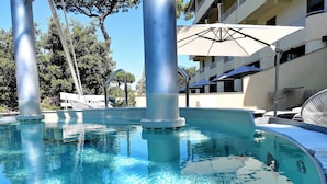 Une piscine aux eaux cristallines, bordée d’une terrasse carrelée, entourée de chaises longues et de parasols, avec un bâtiment visible au loin.
