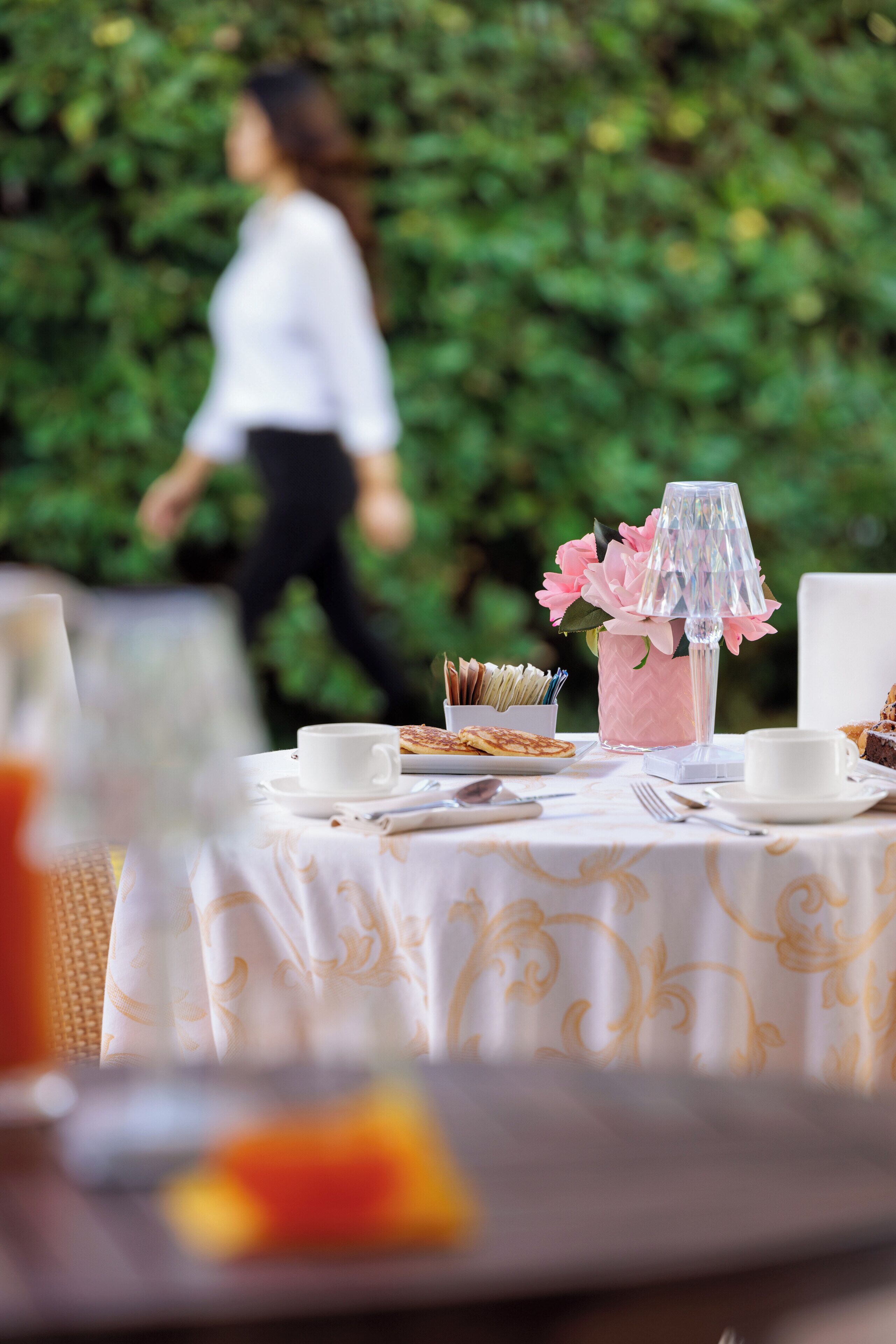 Una mesa cubierta con un mantel blanco, un jarrón rosa con flores y una lámpara de vidrio transparente.