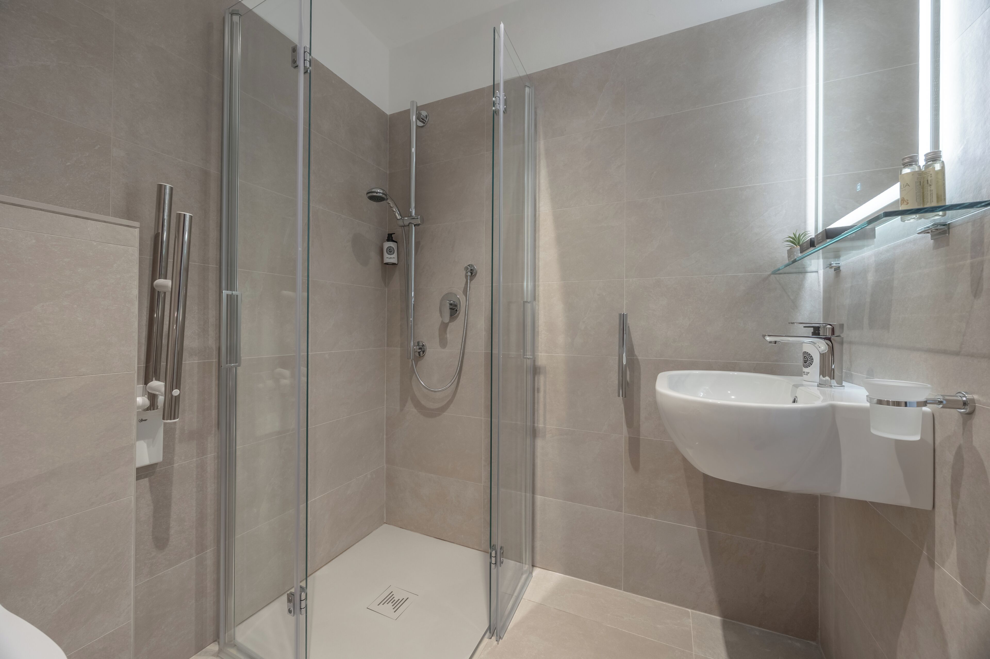 Un baño moderno con ducha envuelta en vidrio, lavamanos blanco y repisa de vidrio con artículos de aseo.