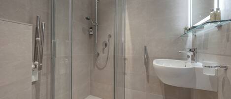 Un baño moderno con ducha envuelta en vidrio, lavamanos blanco y repisa de vidrio con artículos de aseo.