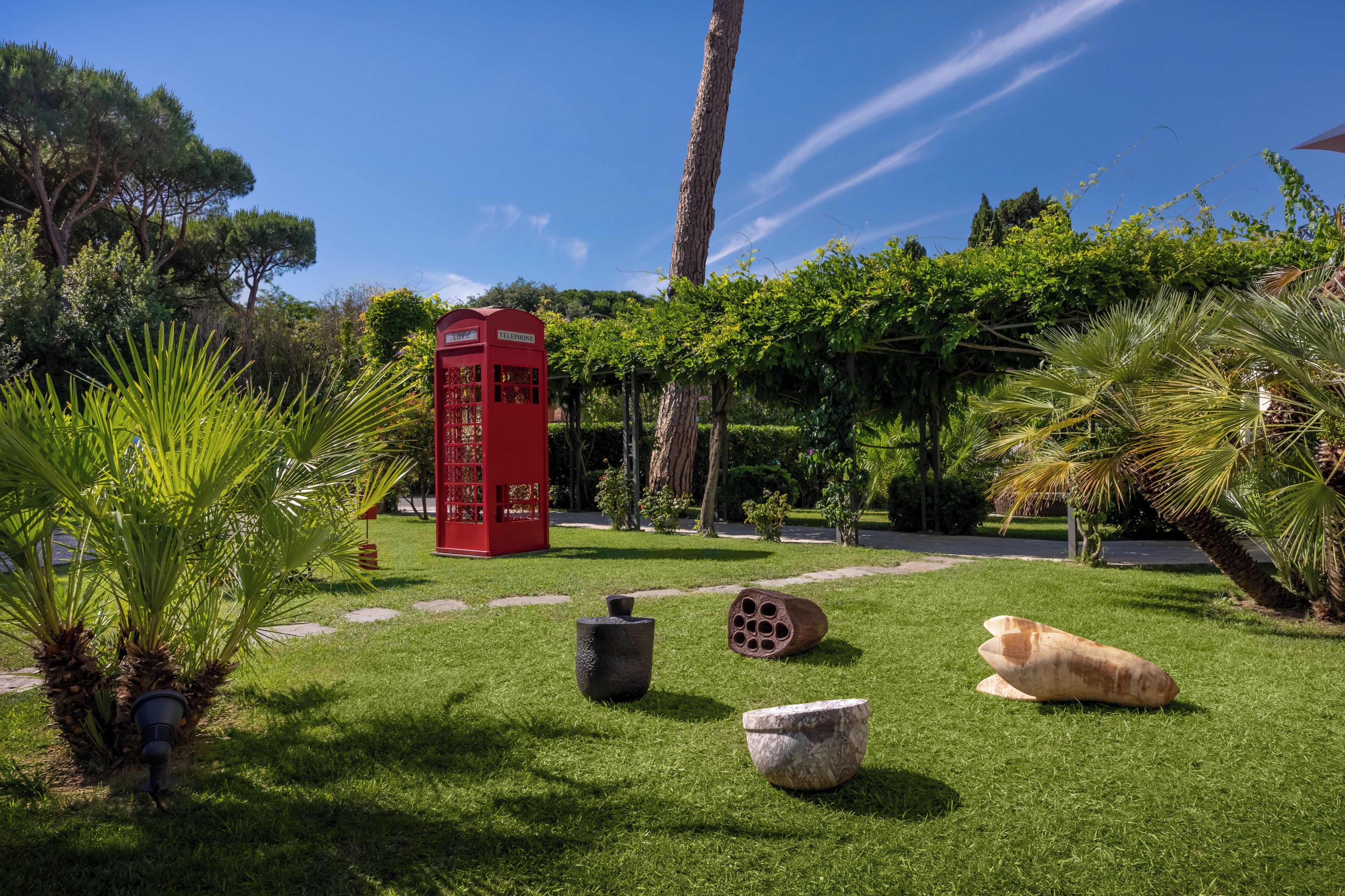 Um jardim com uma cabine telefônica vermelha, vasos de pedra e palmeiras.
