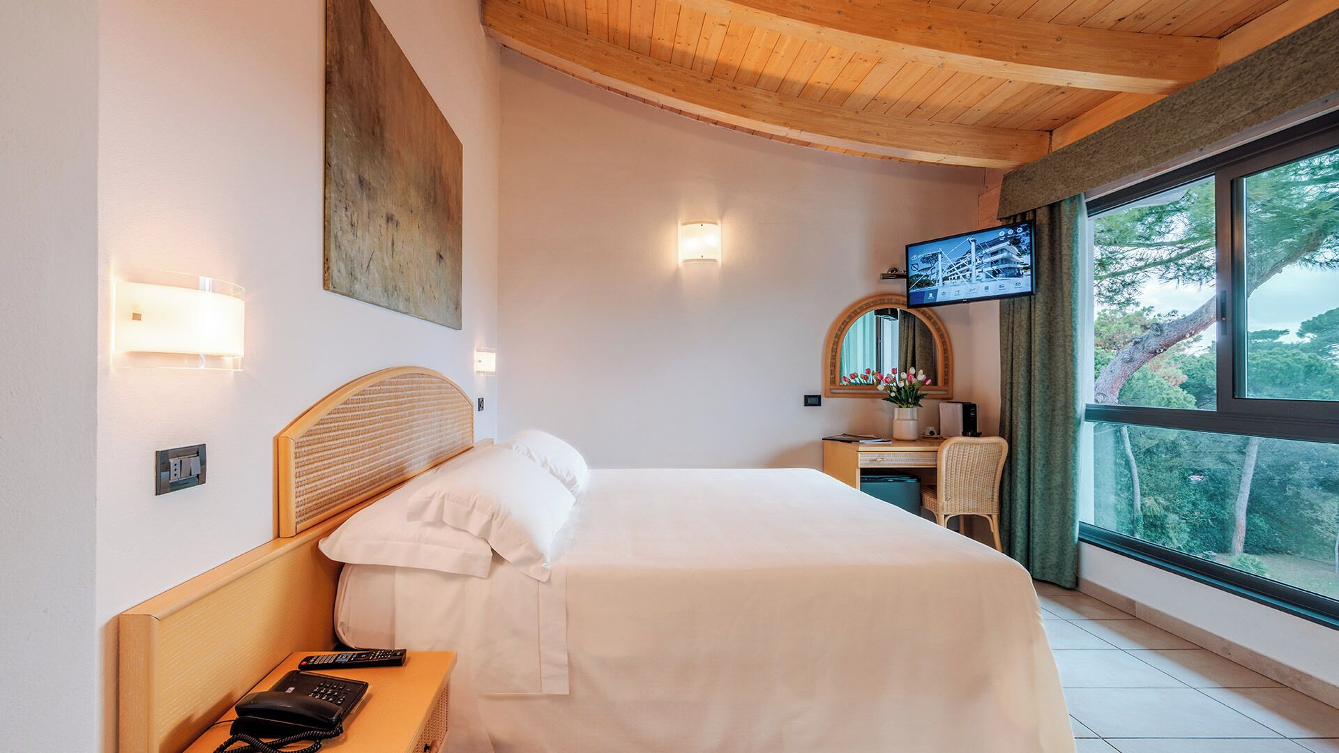 Ein Hotelzimmer mit Bett, Nachttisch, Lampe, Fernseher und einem Fenster mit Blick auf Bäume.