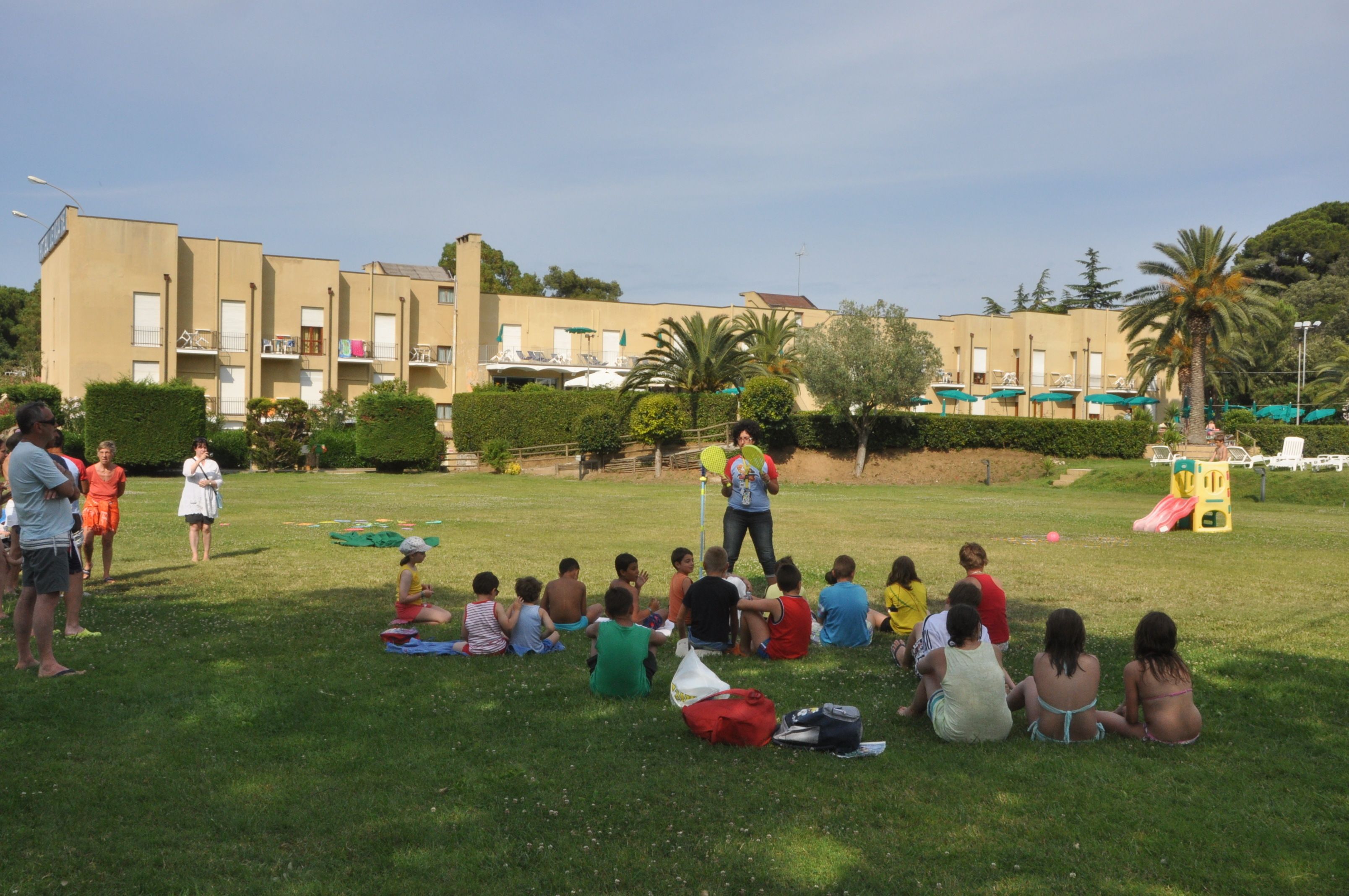 A group of children sitting on grass in a park with a playground in the background.