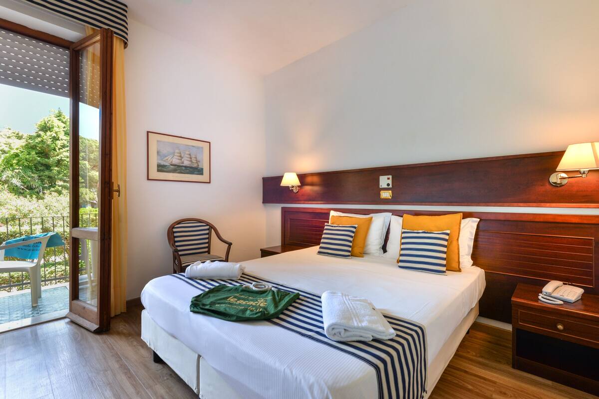 A hotel room with a large bed, a wooden headboard, a desk with a lamp, a chair, and a view of the outdoors through a sliding door.