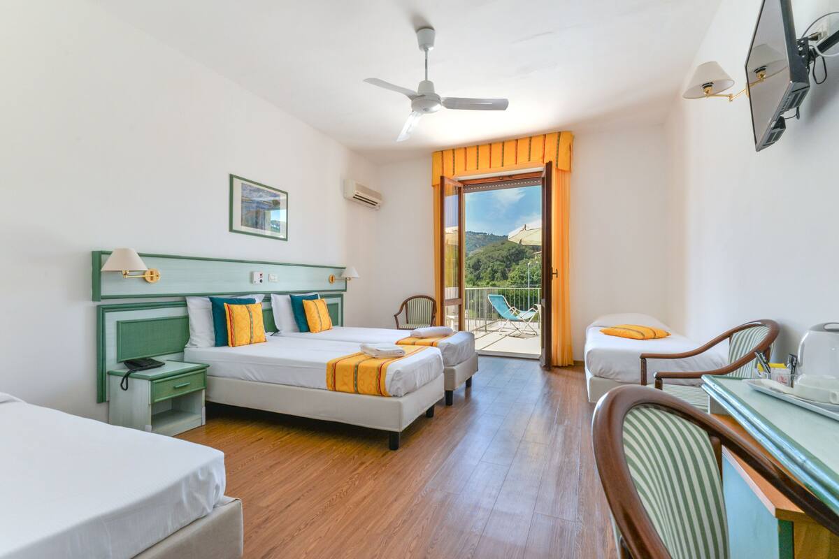 A hotel room with two beds, a desk, a chair, a ceiling fan, and a balcony with a view of greenery.