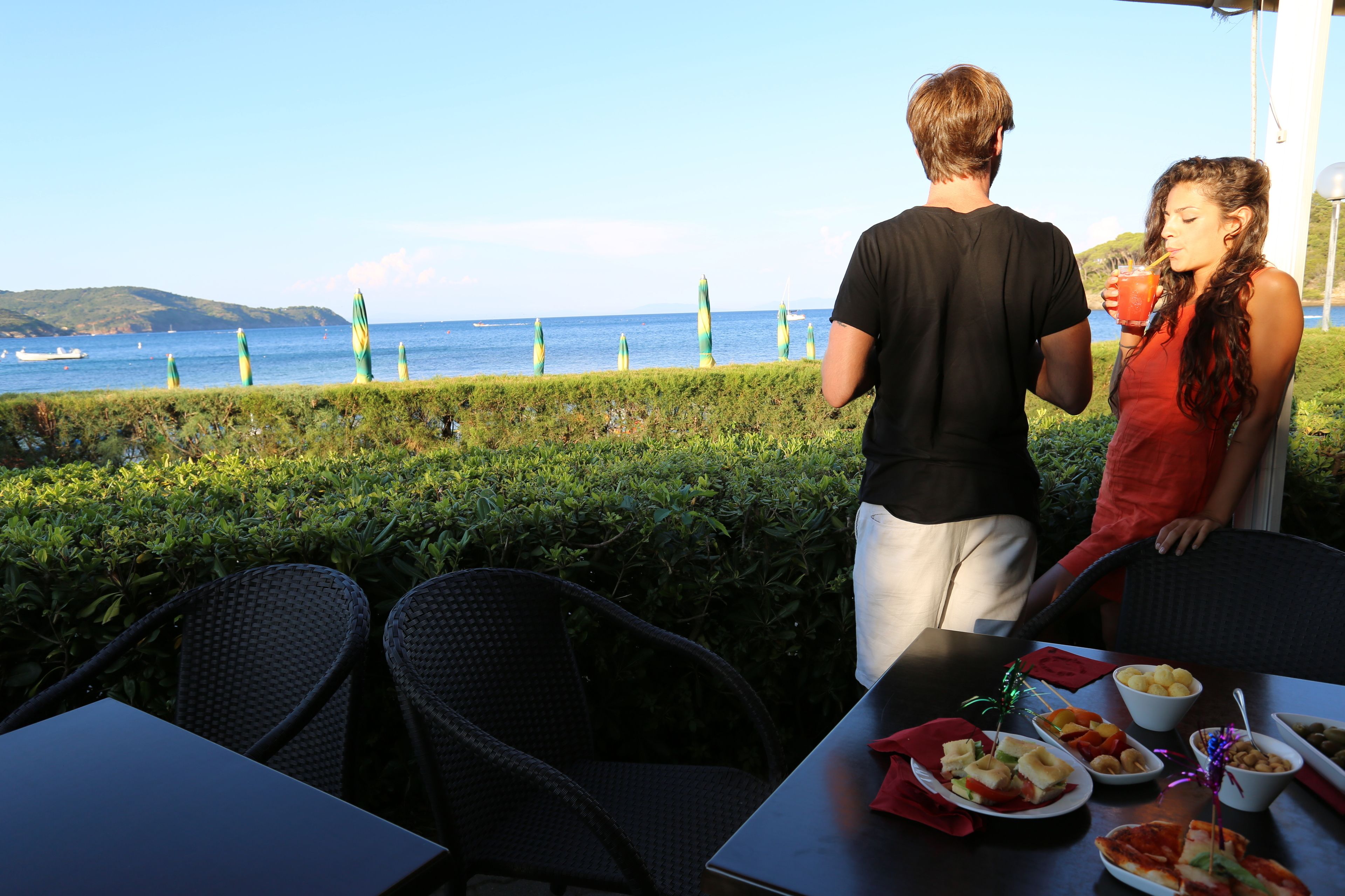 A couple dining at an outdoor table with a view of the sea and distant hills.