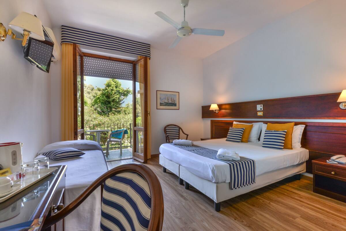 A hotel room with two beds, a balcony, a desk with a chair, and a ceiling fan.