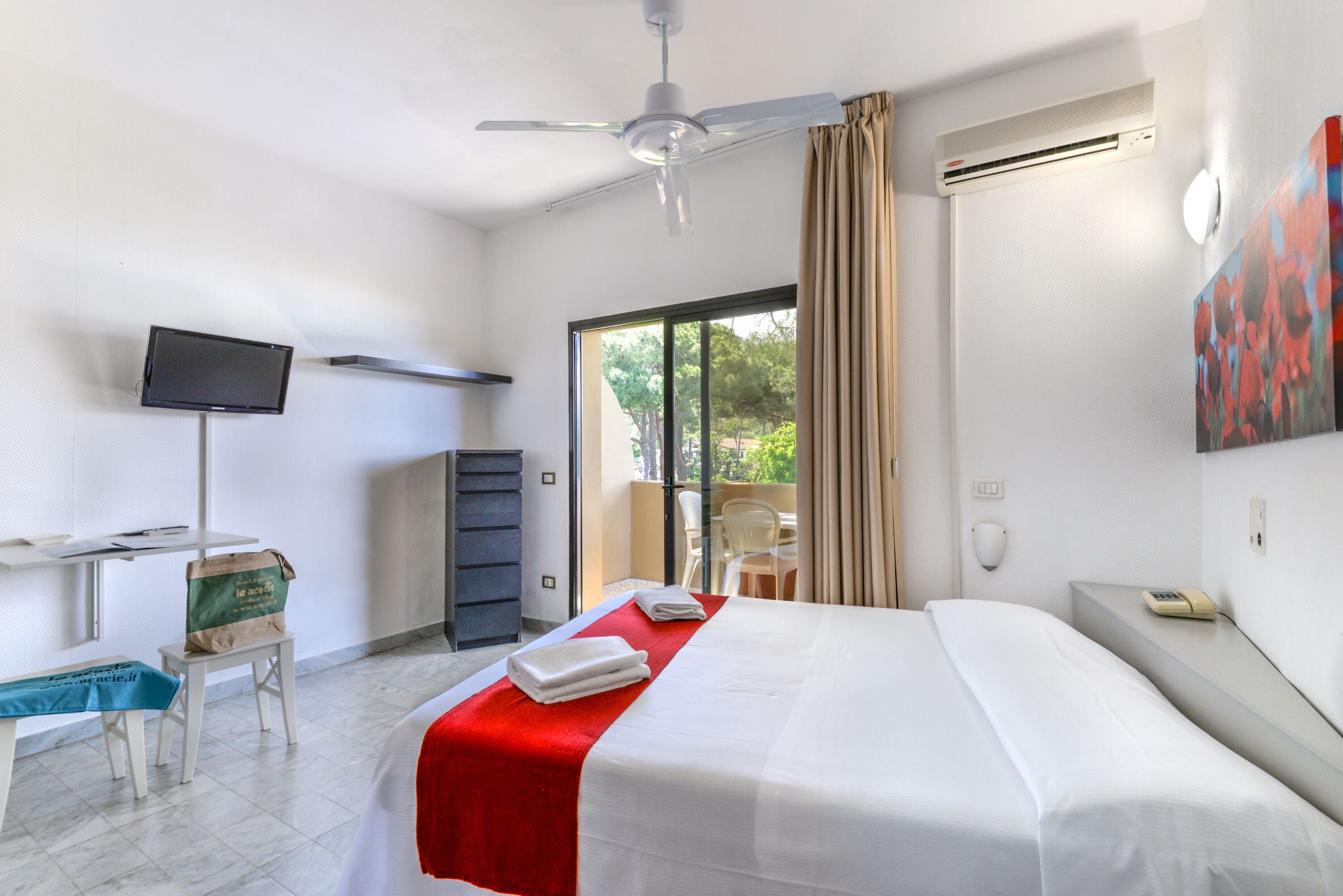 A hotel room with a bed, a desk with a computer, a television, and a sliding glass door to the outside.