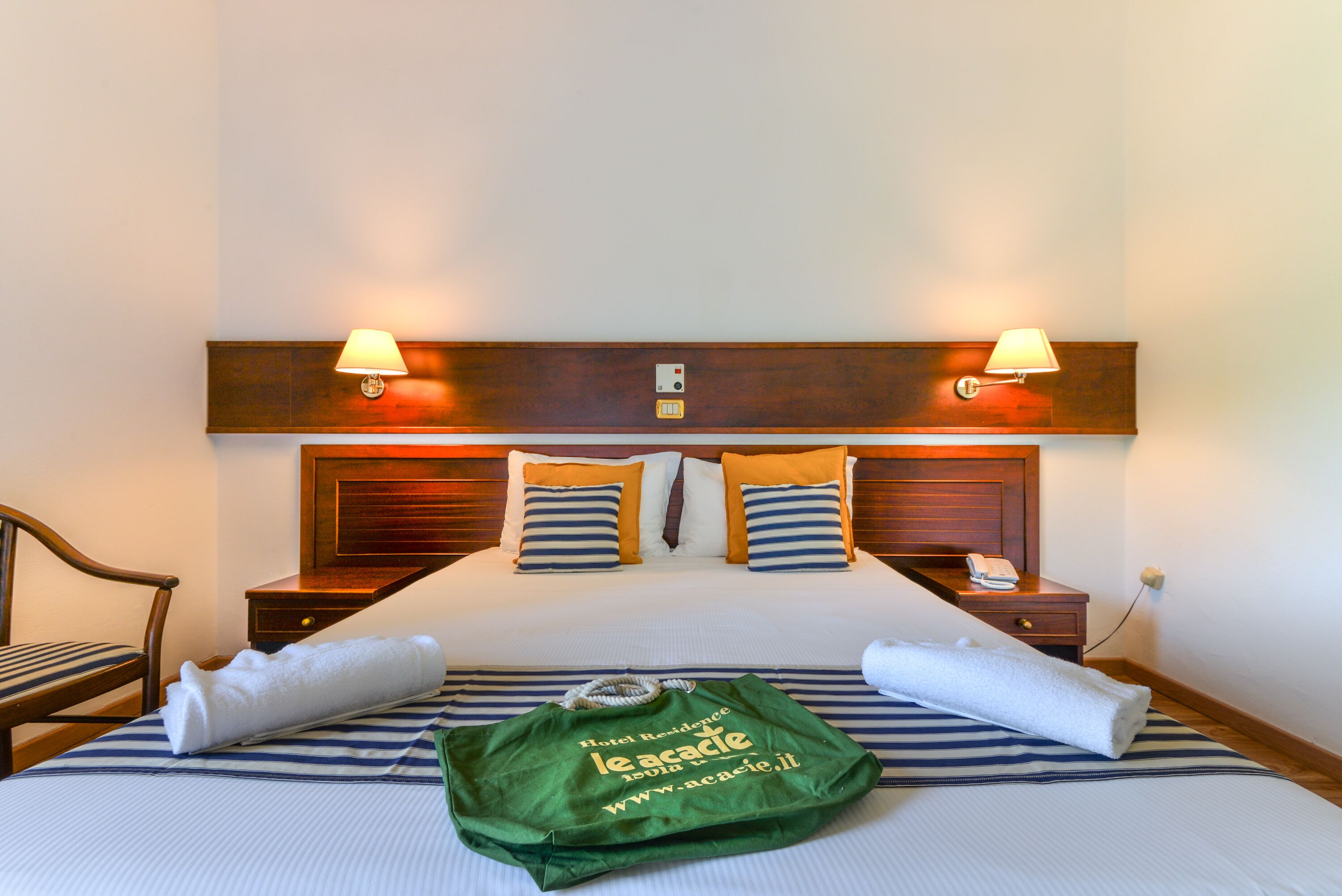 A hotel room with a bed, two bedside lamps, a nightstand, a chair, and a green tote bag on the bed.