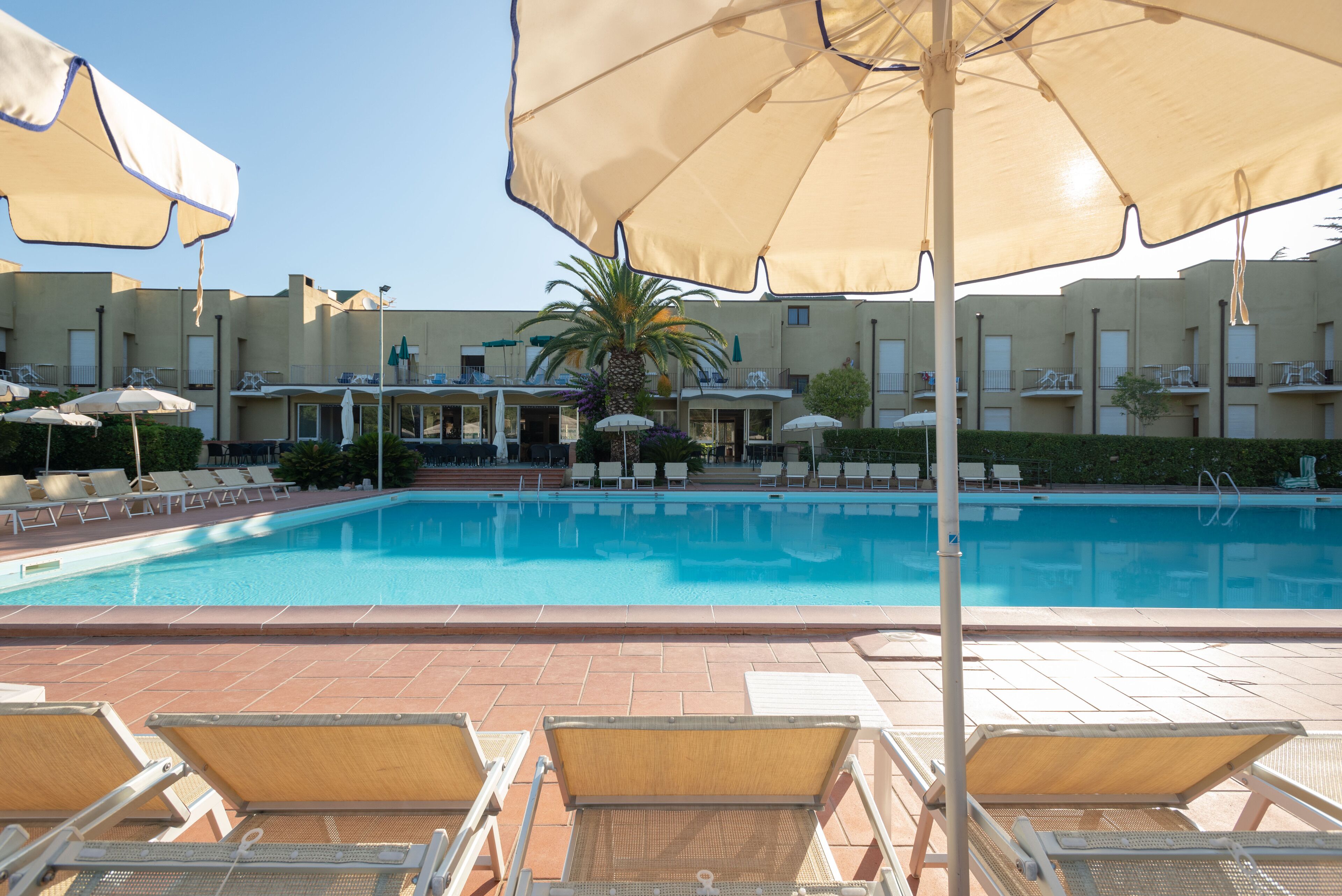 Una piscina con sillones de playa y sombrillas, rodeada por un edificio con varios balcones.