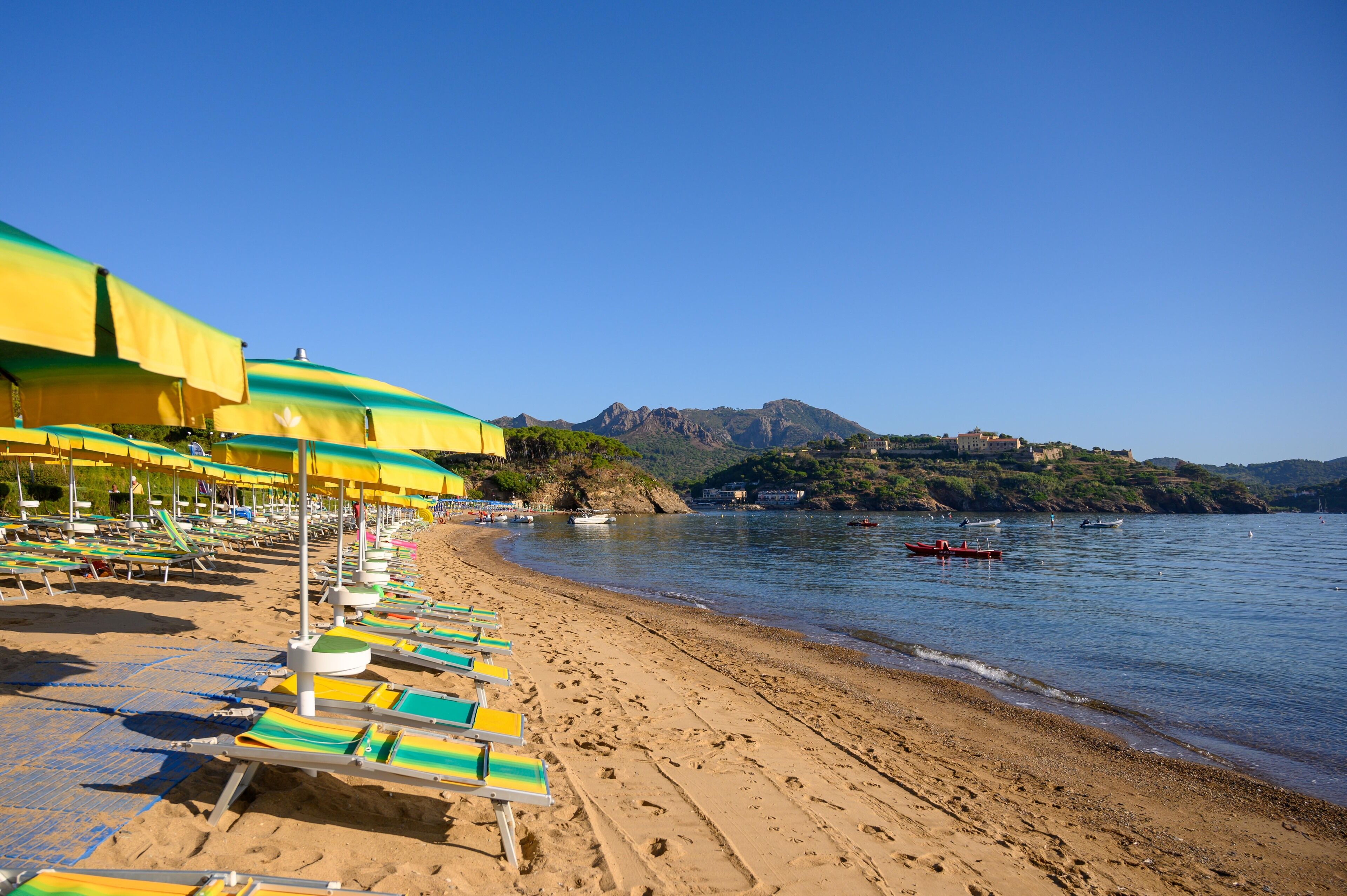 A beach with rows of sun loungers under umbrellas, a calm sea, and a mountainous backdrop.