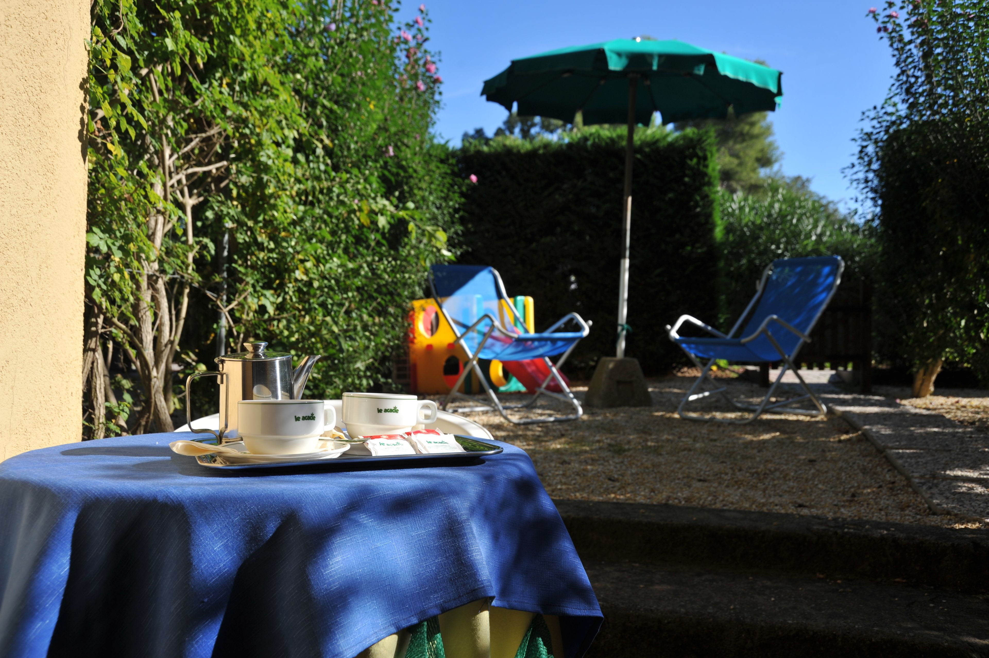 A table with a blue tablecloth, a white teapot, cups, and a tray with food items. There are two blue lounge chairs and a green umbrella in the background.
