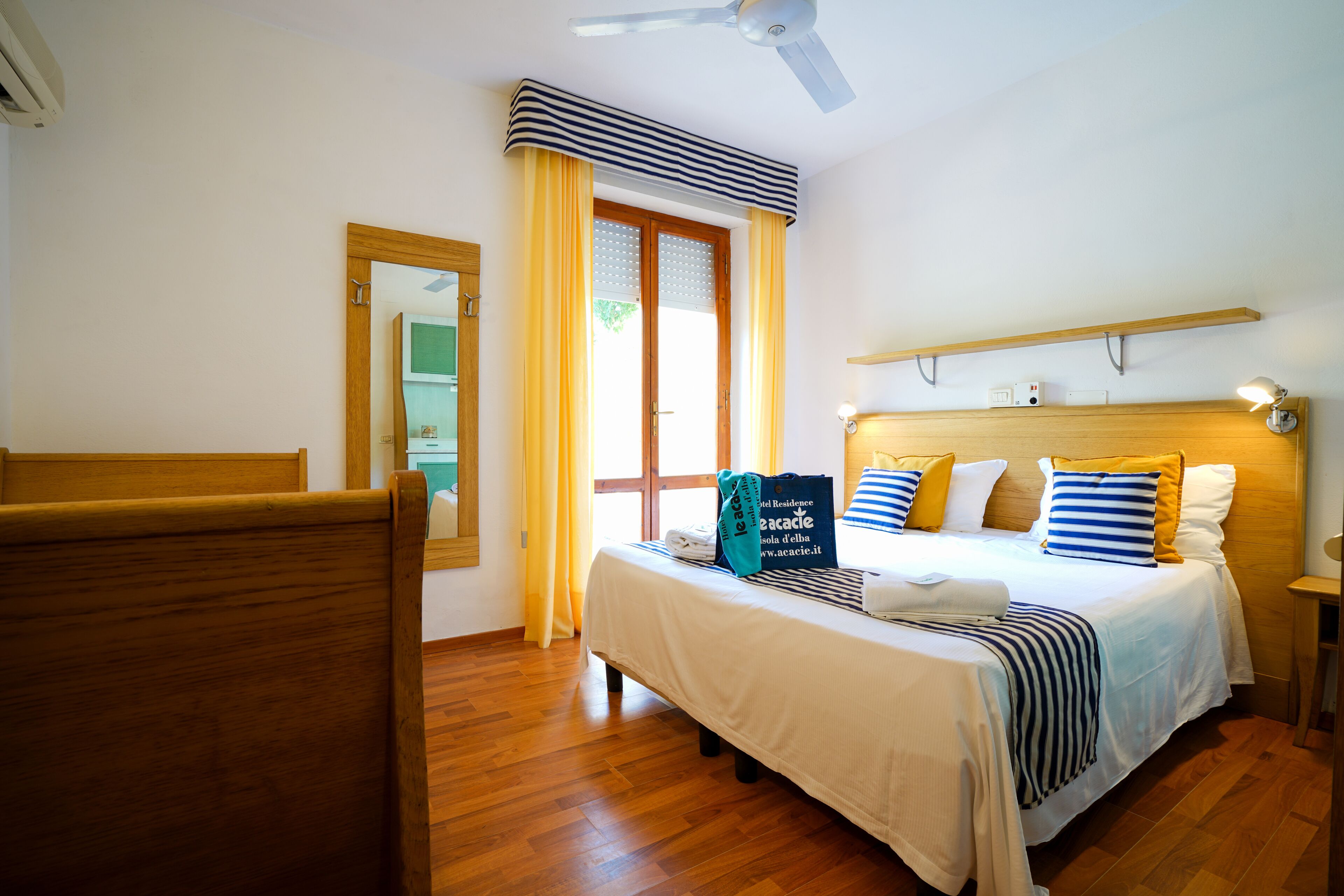 A hotel room with a wooden bed, a ceiling fan, a window with curtains, a bedside table with a lamp, and a wooden shelf above the bed.