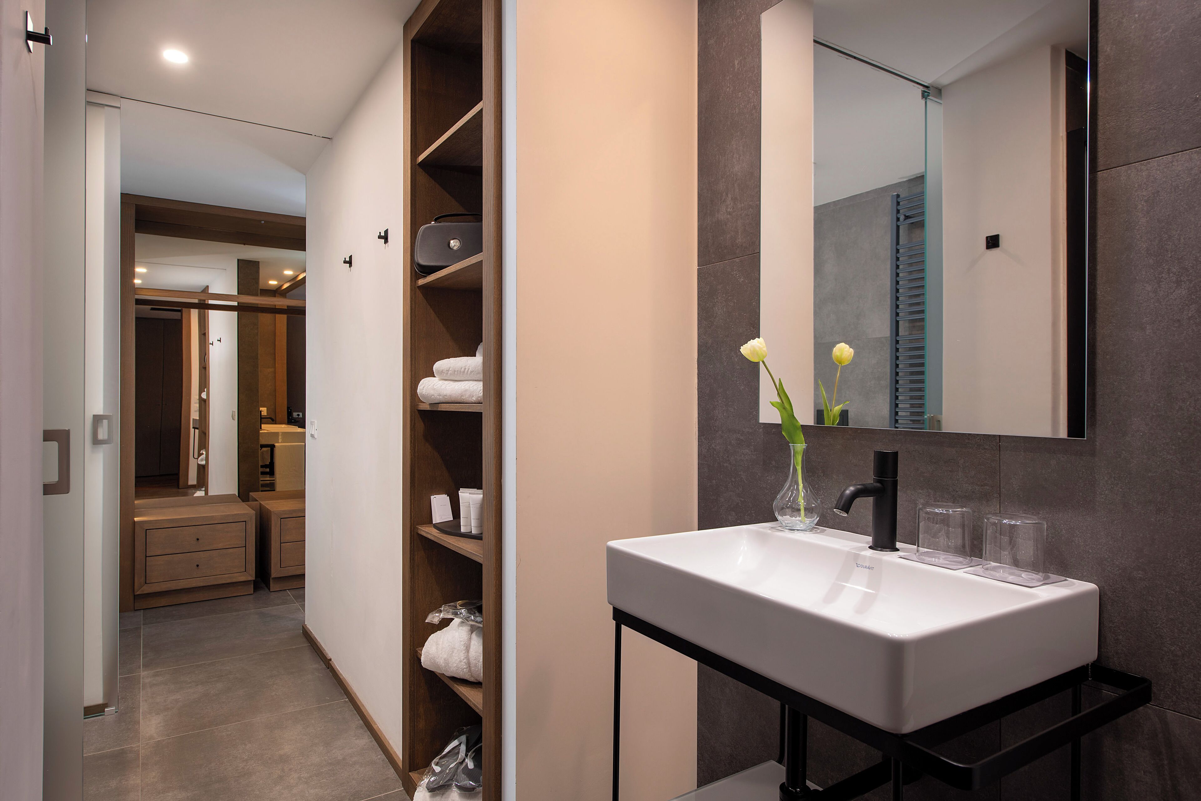 Une salle de bain moderne avec un lavabo blanc, un miroir et un vase rempli de fleurs. On y trouve une étagère en bois où sont posés des serviettes et des articles de toilette.