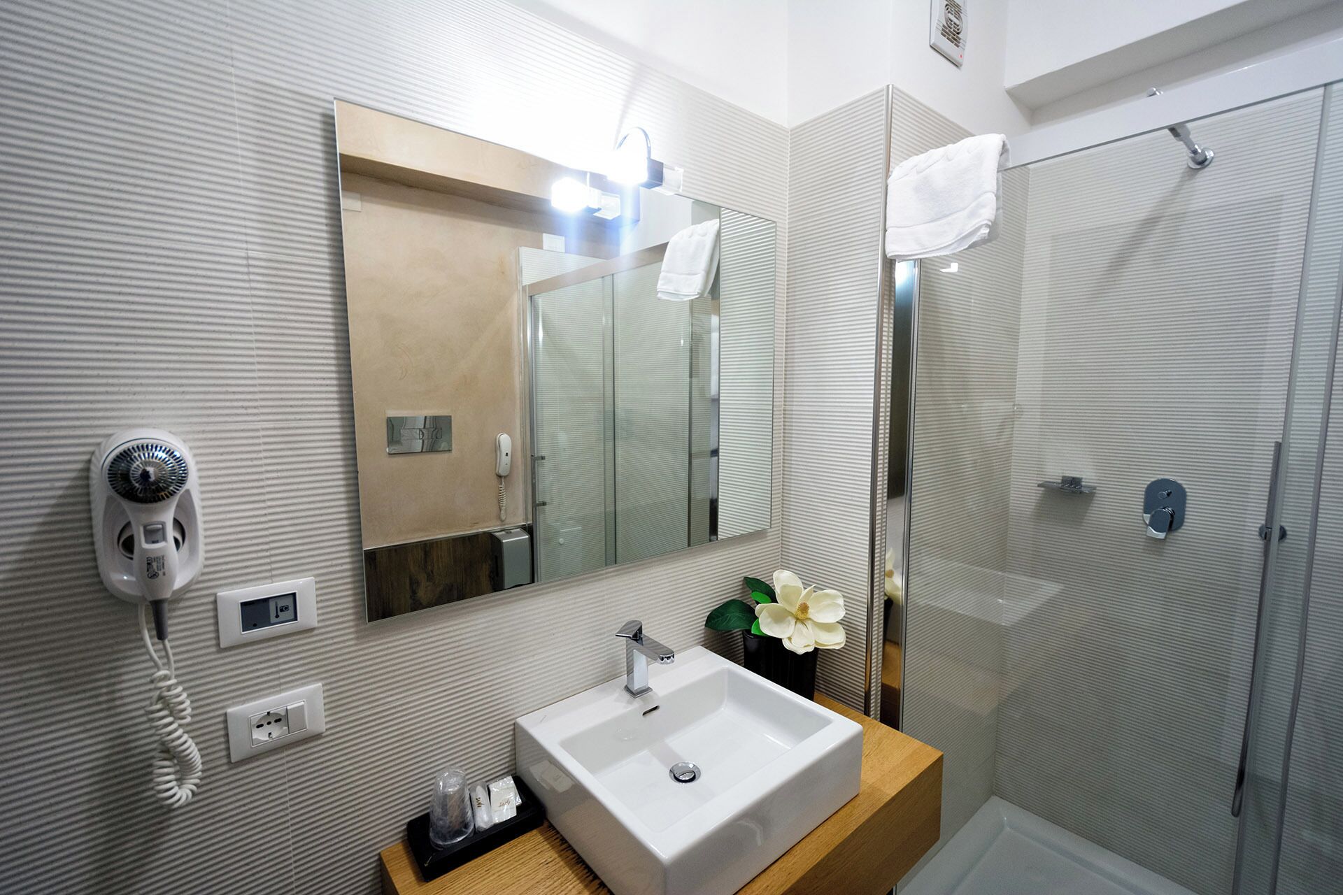 A modern bathroom with a large mirror, a sink, a shower, and a hairdryer mounted on the wall.