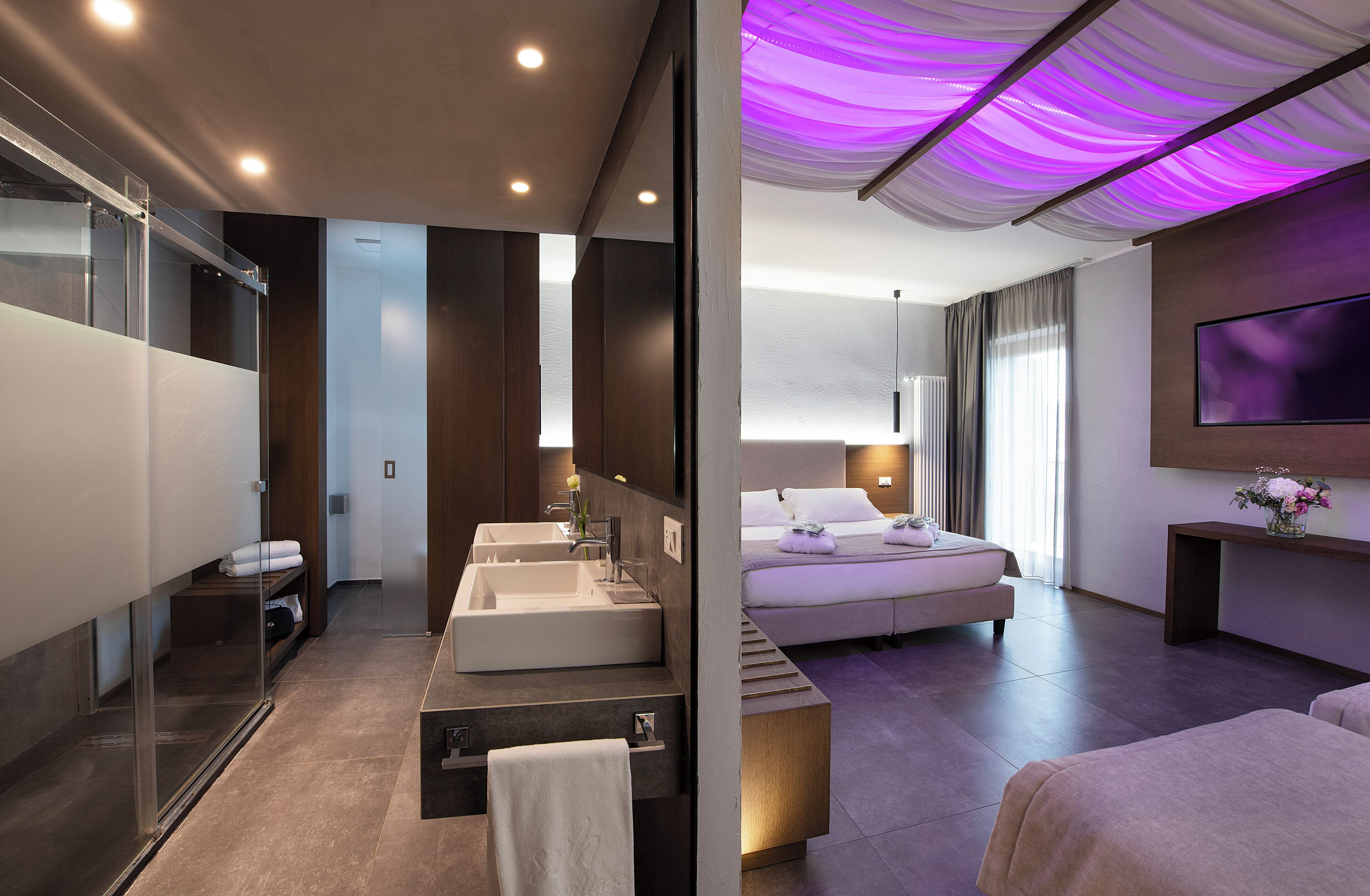 A modern hotel room with a large bed, a separate bathroom with a vanity, a flat-screen TV mounted on the wall, and a large window providing natural light.