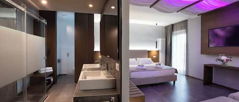 Una habitación de hotel moderna con una cama grande, baño independiente con tocador, televisor de pantalla plana montado en la pared y un amplio ventanal que proporciona luz natural.