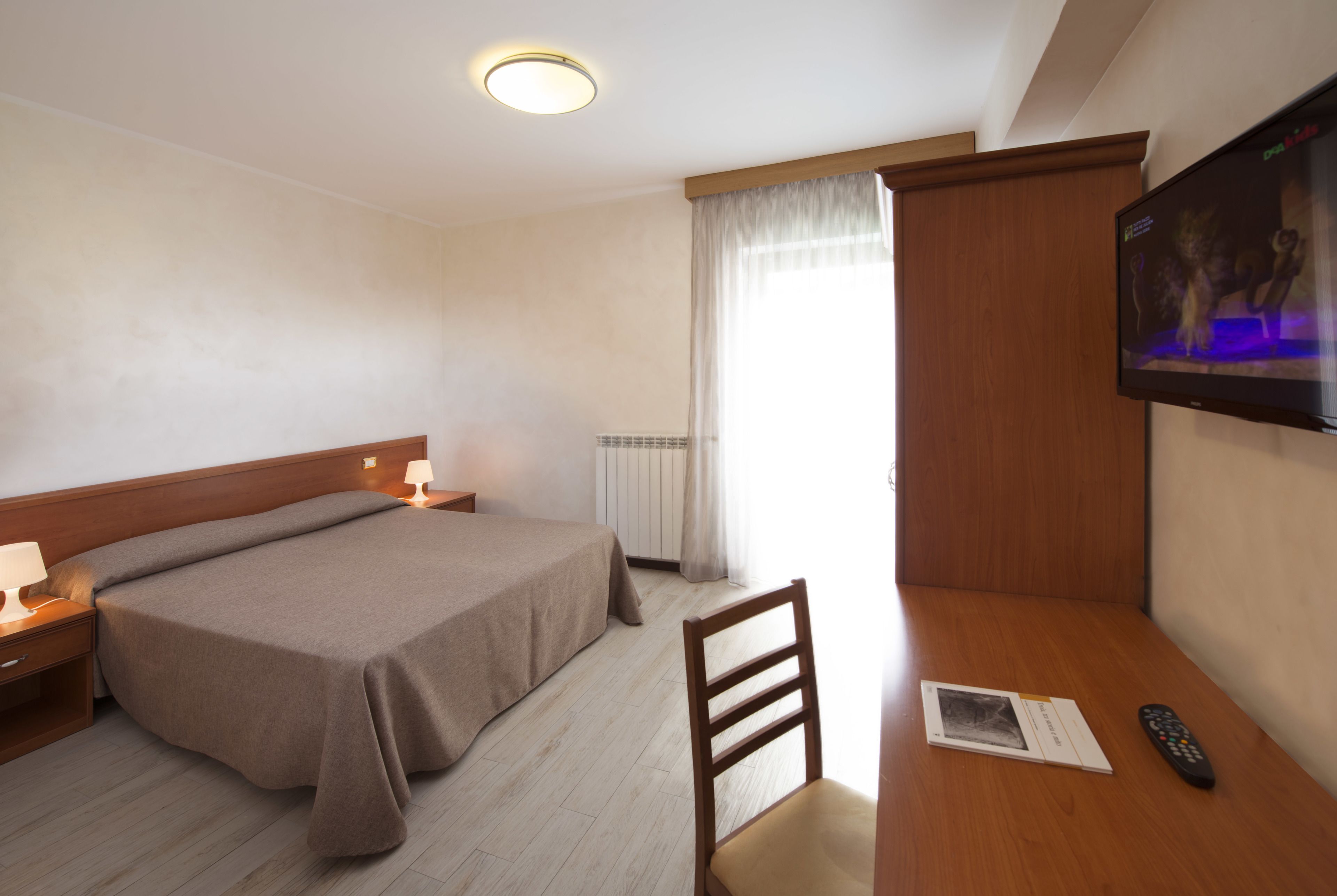 A hotel room with a bed, bedside table, lamp, chair, TV, and a remote control on a wooden desk.