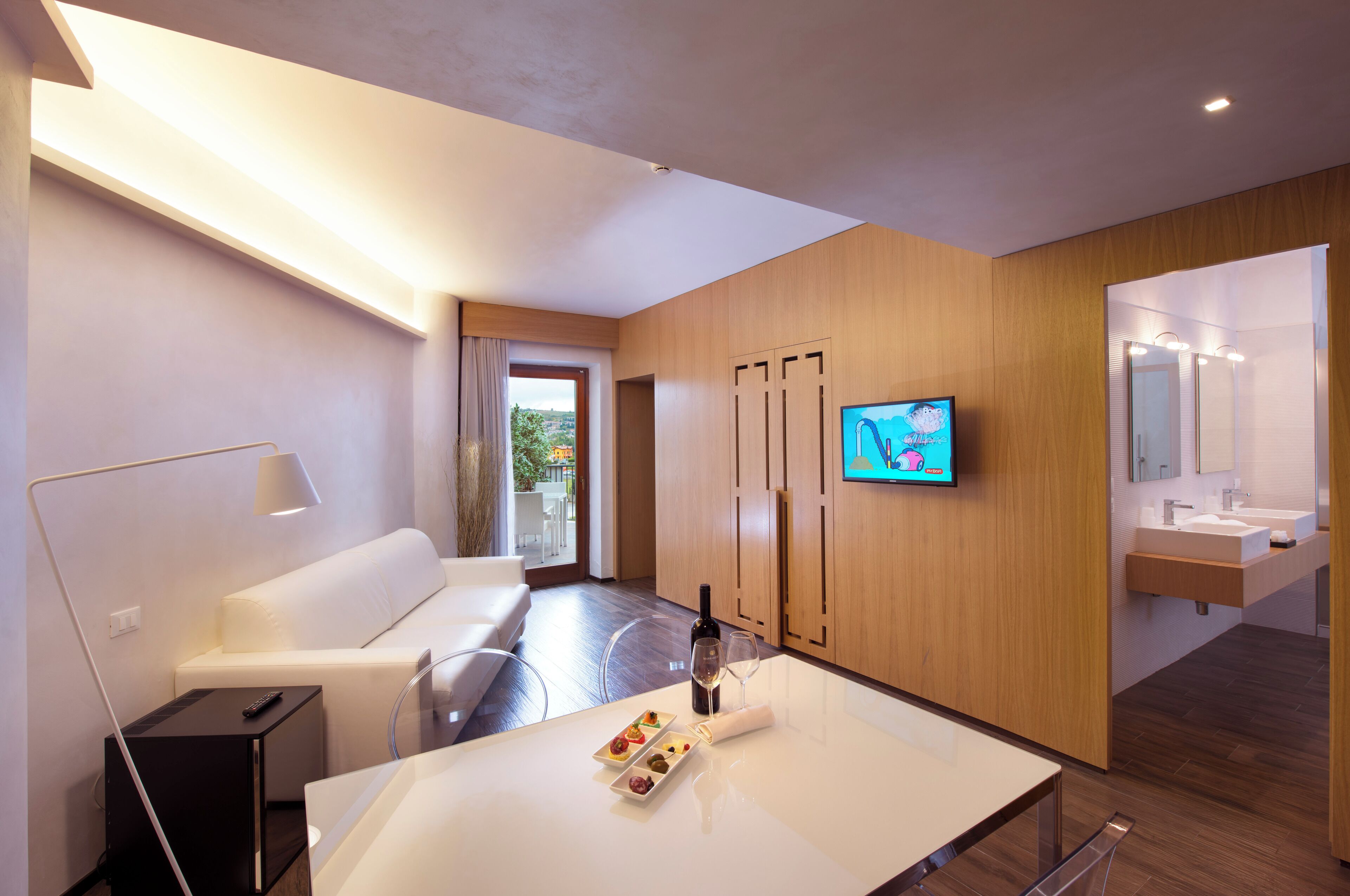 A modern hotel room with a white sofa, a small table with a bottle and glasses, a TV on the wall, and a bathroom with a mirror and sink.