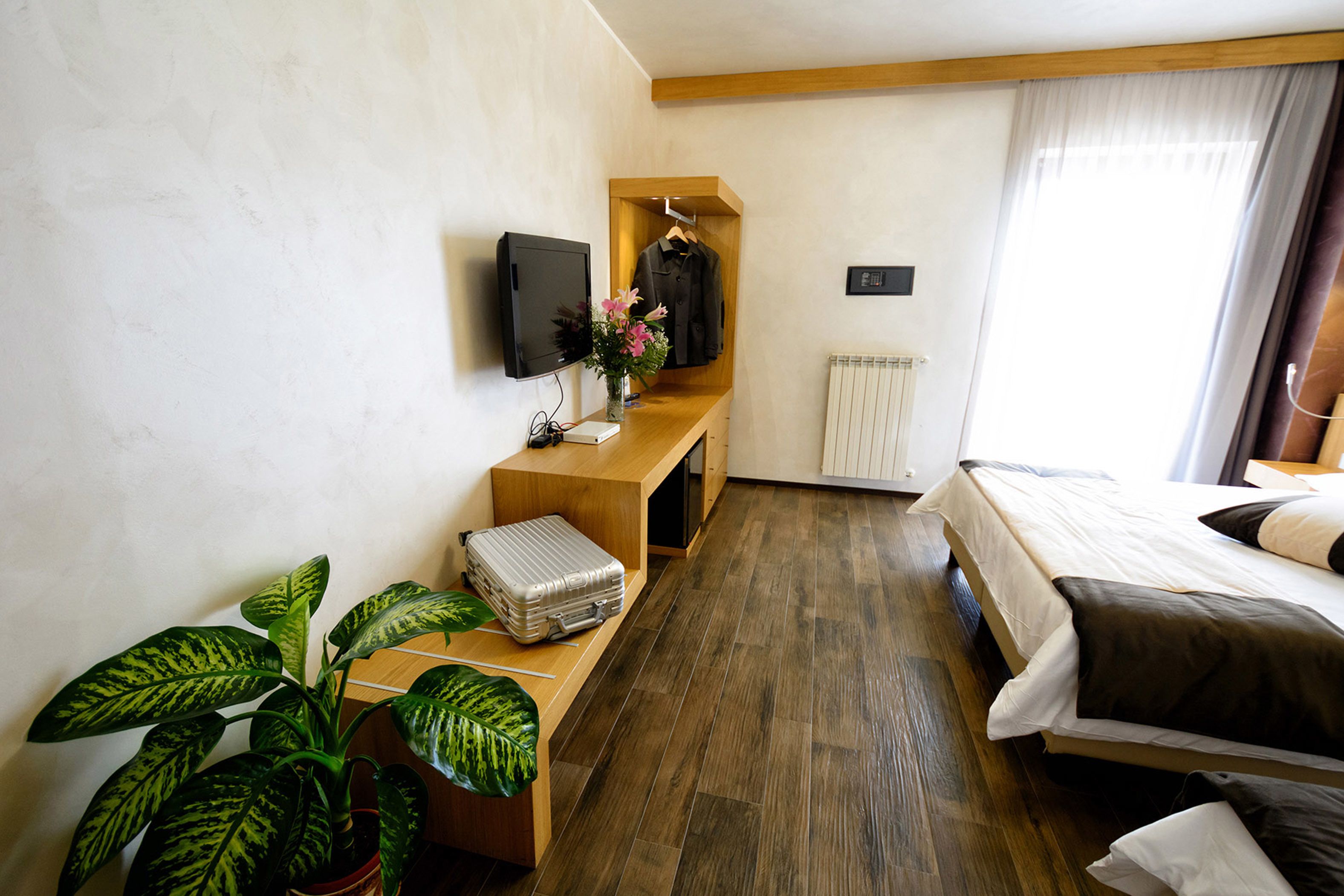 Una habitaci ón de hotel con una cama de madera, una mesita de noche con una maleta, un televisor, un perchero y una planta en maceta.
