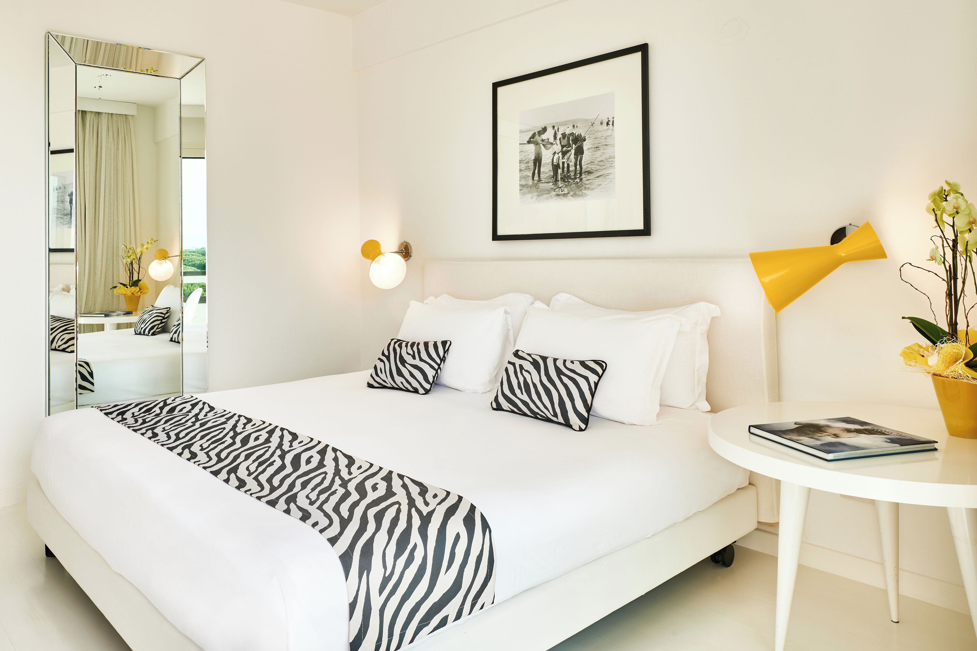 A hotel room with a large bed, a bedside table with a lamp, a framed picture on the wall, and a mirror.