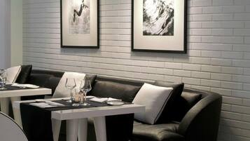 Un espace repas moderne avec un canapé en cuir noir, des tables blanches et des luminaires suspendus noirs. Deux photographies en noir et blanc, encadrées, sont accrochées au mur.