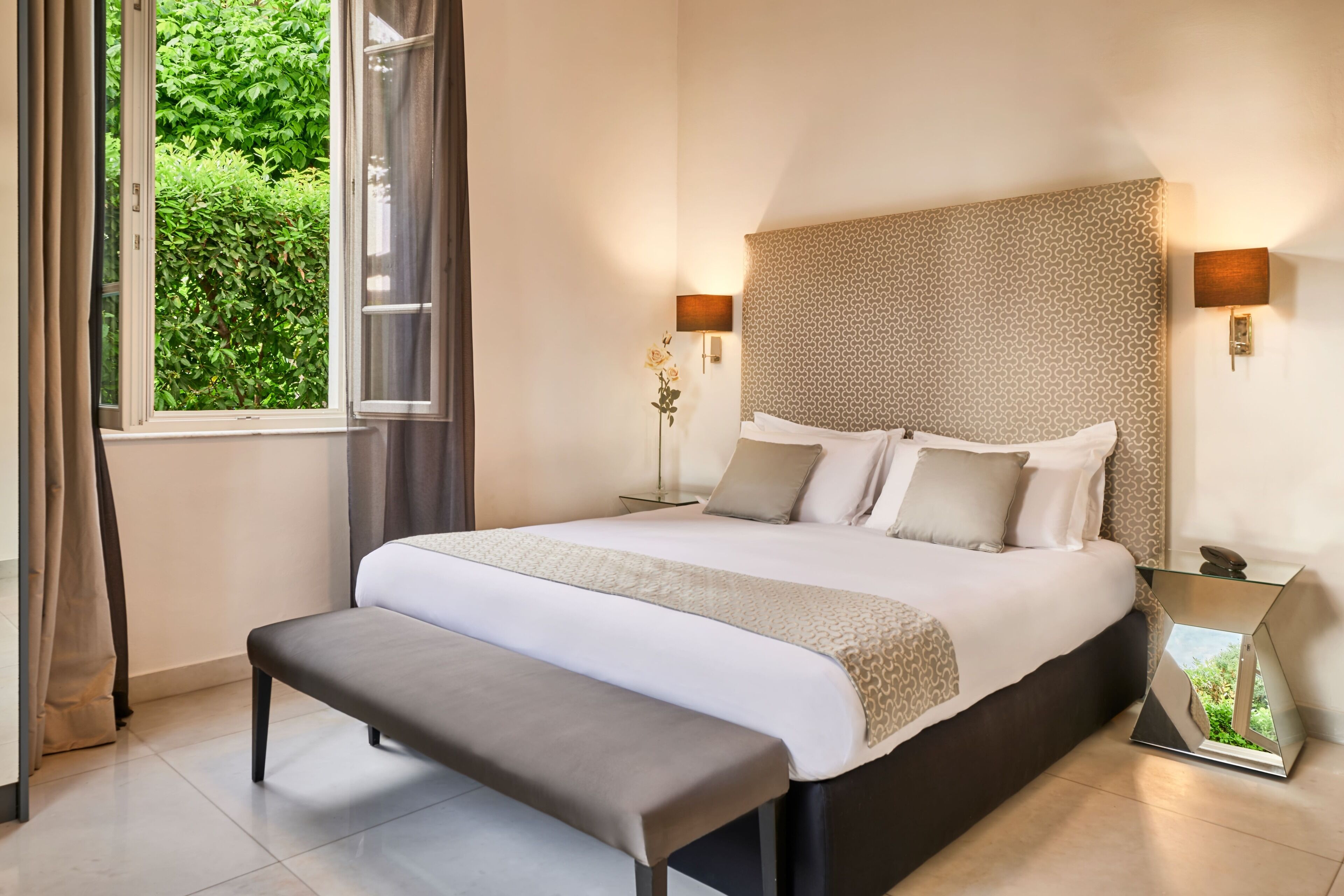 Een keurig ingerichte slaapkamer met een groot bed, een bankje, een nachtkastje en een raam met gordijnen.