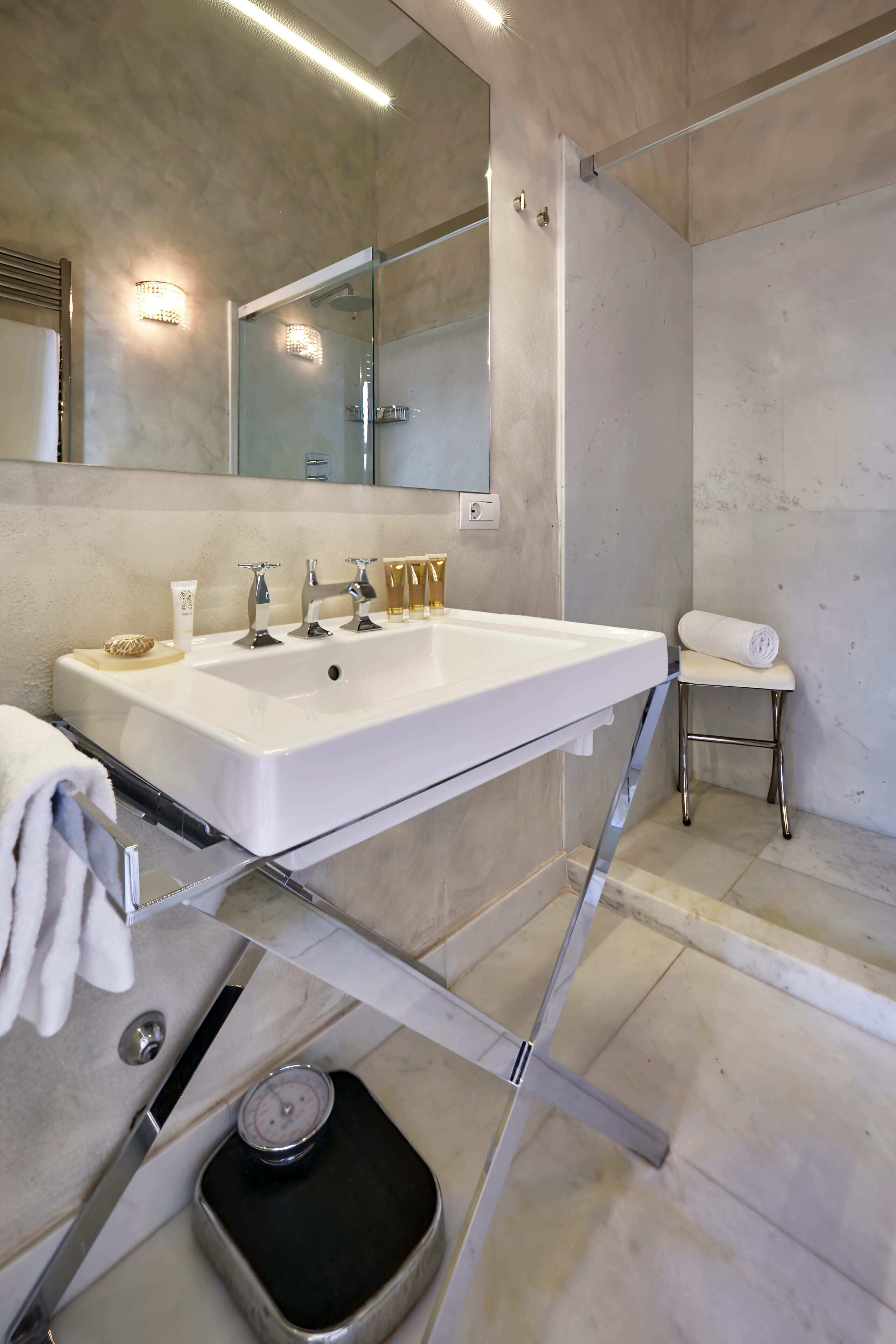 Une salle de bain moderne avec un grand lavabo blanc, un miroir, une douche et un petit tabouret sur lequel est posée une serviette.