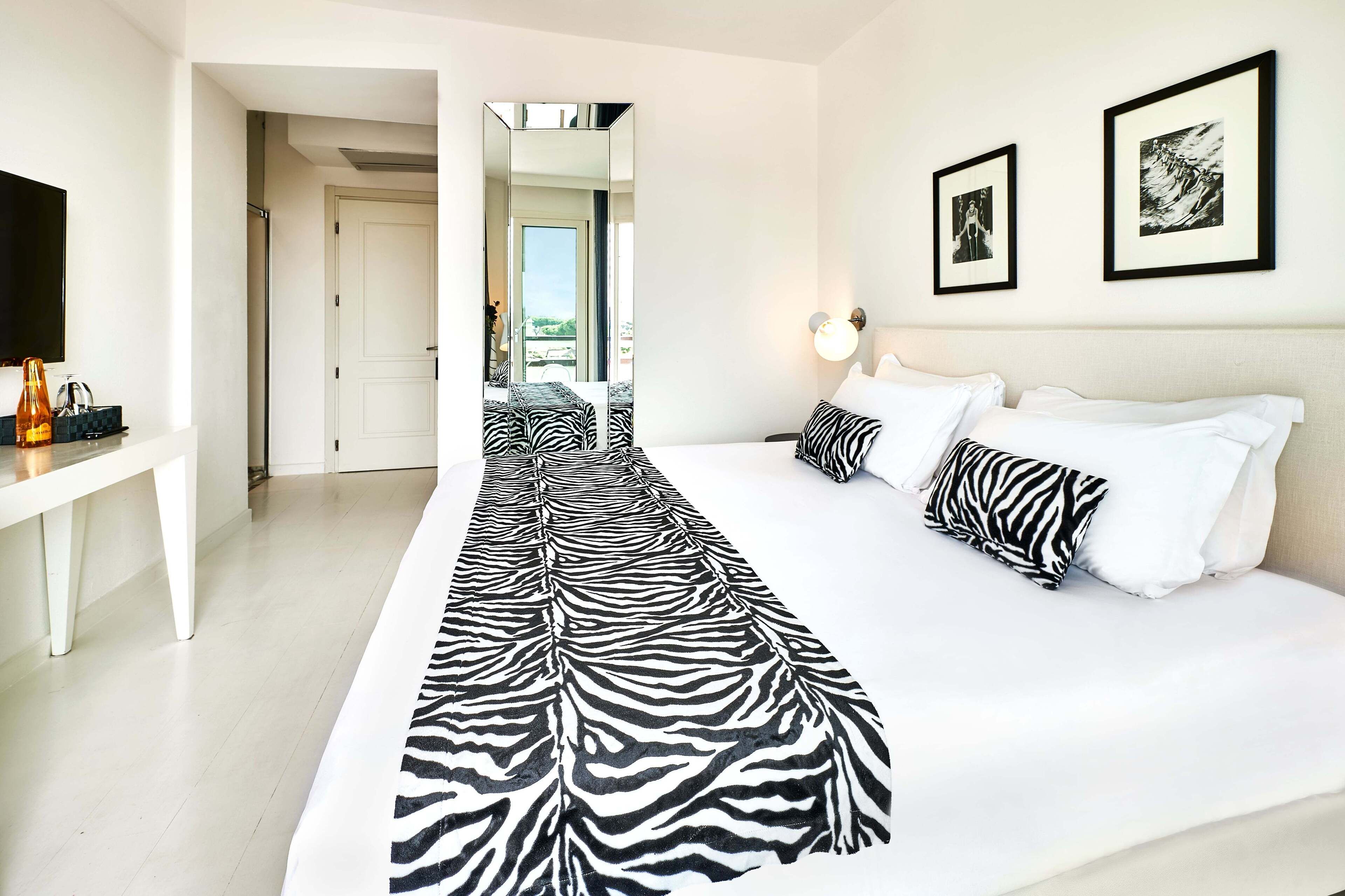 A hotel room with a zebra-patterned rug, a bed with white bedding, a desk with a flat-screen TV, and framed artwork on the wall.