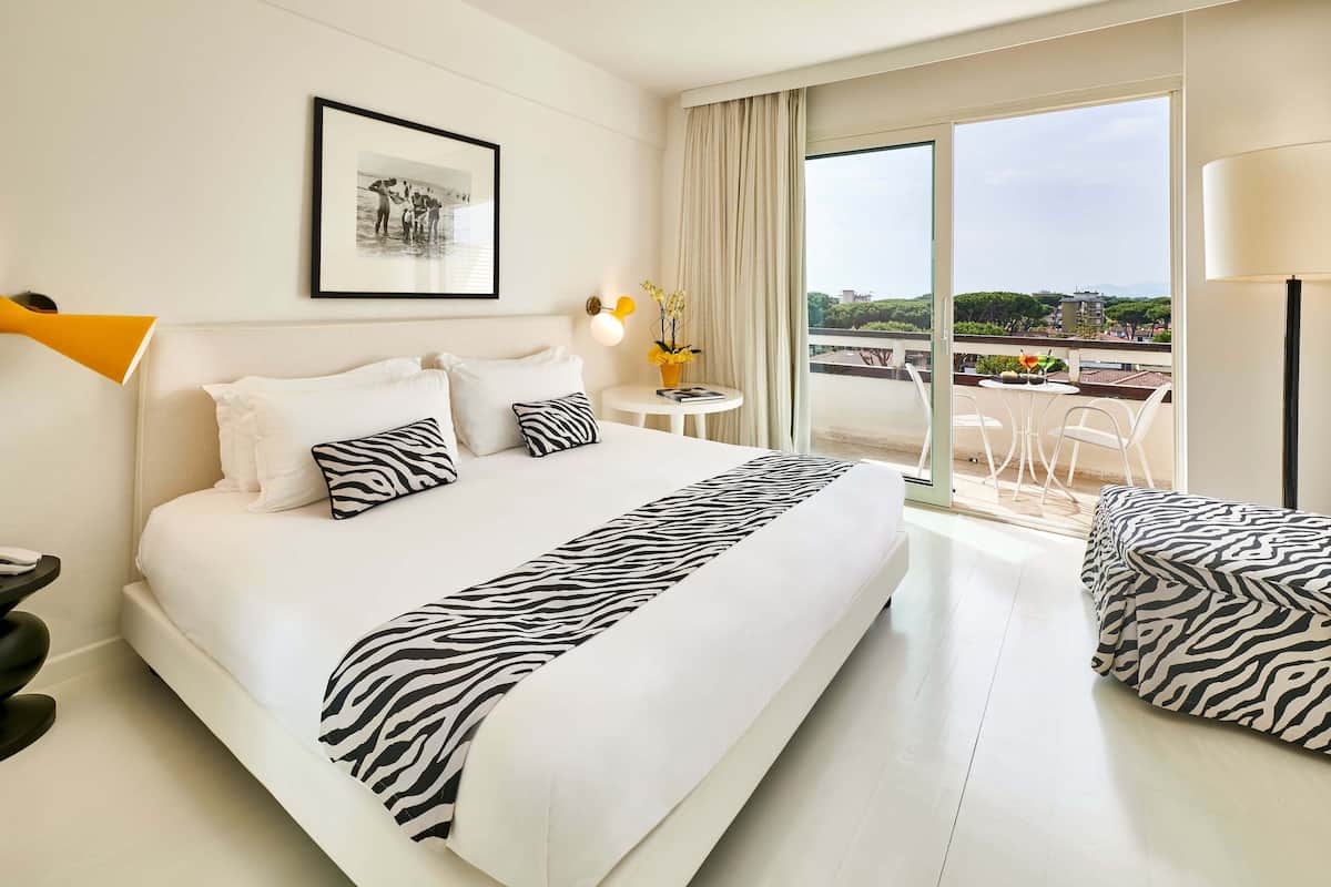 Hotellihuone, jossa on suuri sänky, pieni pöytä lampun kanssa, seinällä kehystetty taulu ja parveke, jolta on näkymä.