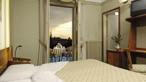 A hotel room with a bed, a bedside table with a lamp, a small table with chairs outside the door, and a view of the sky. - Cacciani Hotel (Frascati)