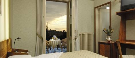 Habitación de hotel con cama, mesita de noche con lámpara, pequeña mesa con sillas afuera de la puerta y vista al cielo.