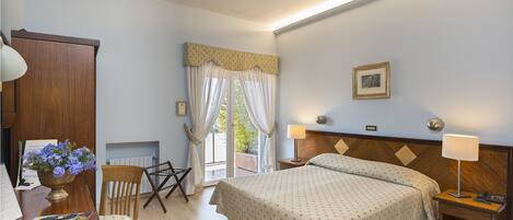 A hotel room with a bed, bedside tables, a desk with a chair, a window with curtains, and a framed picture on the wall.