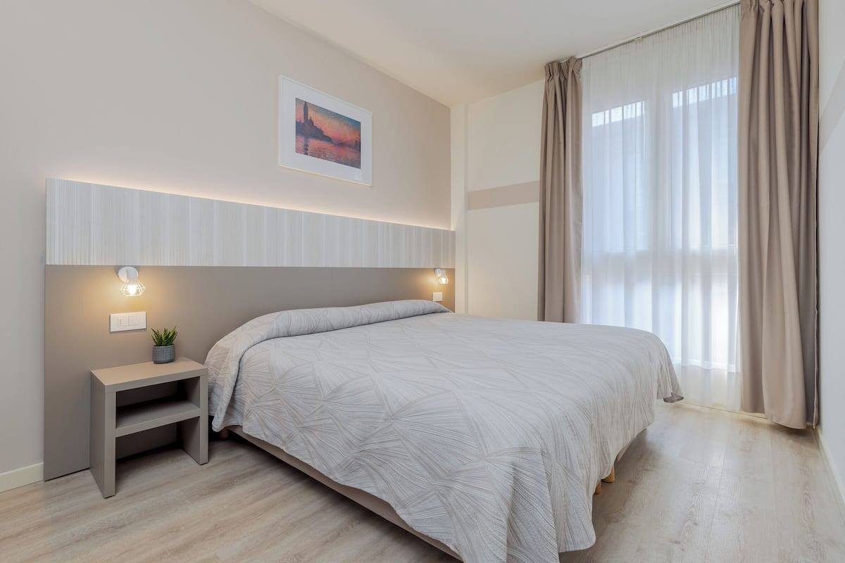 A modern bedroom with a bed, bedside tables, a wall-mounted light, and a window with curtains.