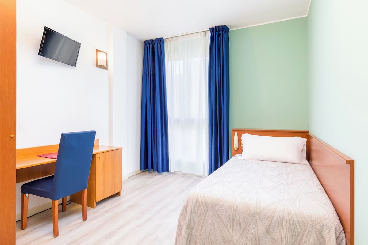 A hotel room with a bed, a desk with a chair, a TV mounted on the wall, and a window with curtains.