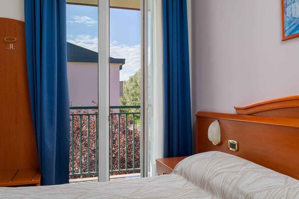A hotel room with a bed, a wooden headboard, a nightstand, and a view of a balcony with a railing and trees.