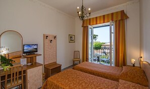 A hotel room with two beds, a television, a desk with a computer, a chair, a small table with a plant, and a balcony with outdoor furniture.