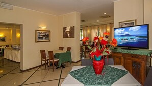 A hotel room with a dining area, a television displaying a scenic ocean view, and a table with a red vase of flowers. - Hotel Villa Ombrosa (Pietrasanta)