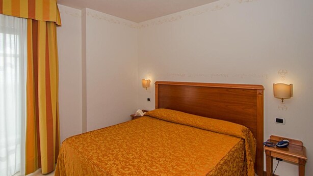 A hotel room with a wooden bed, two wall-mounted lamps, a nightstand, and a striped curtain. - Hotel Villa Ombrosa (Pietrasanta)