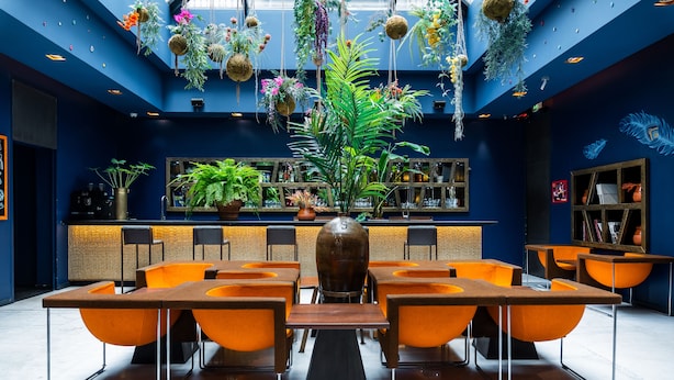 A modern restaurant with orange chairs and a central table, surrounded by potted plants and hanging decorations. - 1K Paris (Paris)