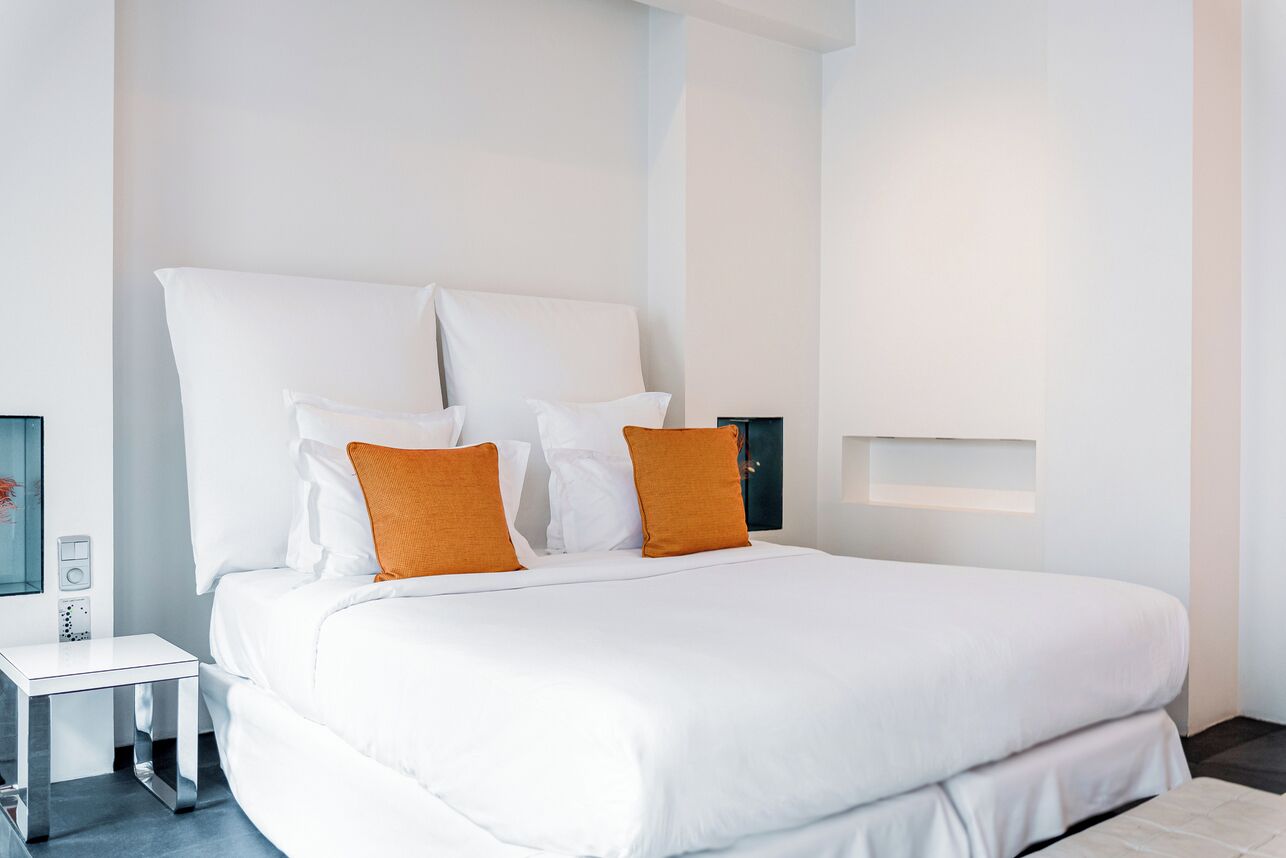 A modern hotel room with a large bed, white bedding, and two orange pillows.