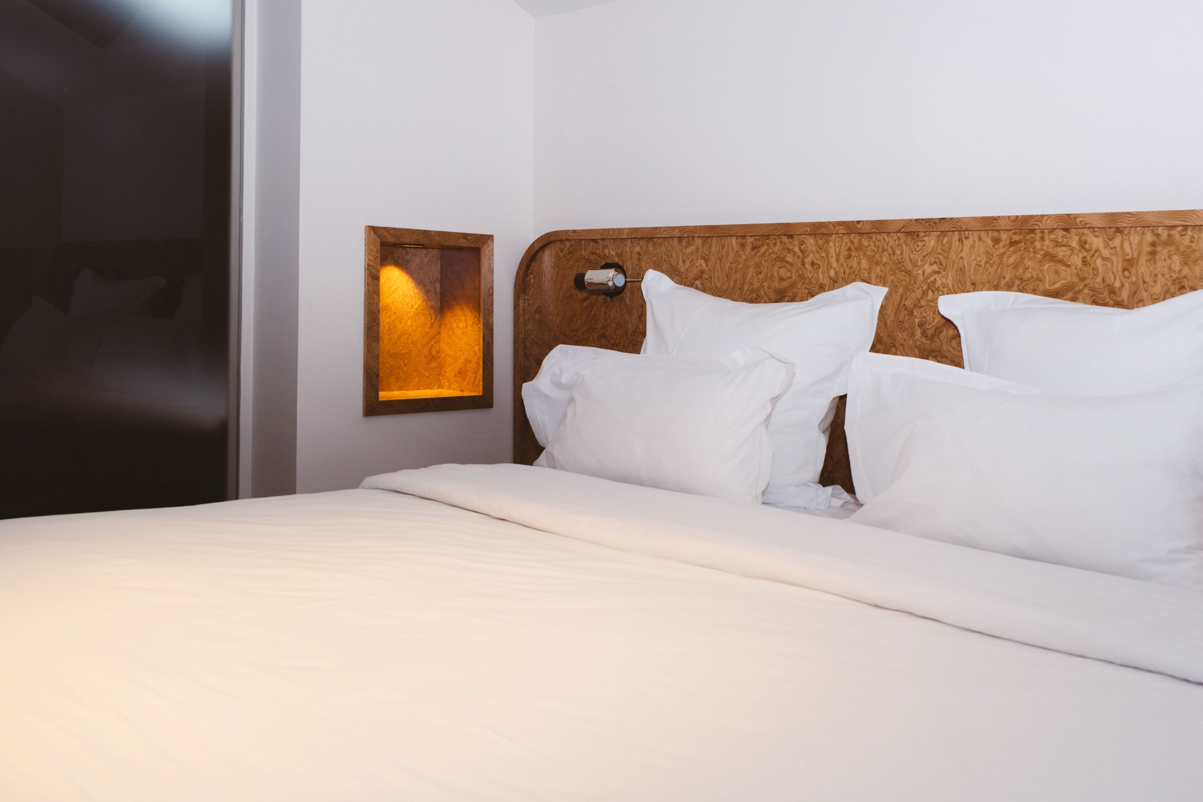 A neatly made bed with white linens, a wooden headboard, and a wall-mounted light fixture.